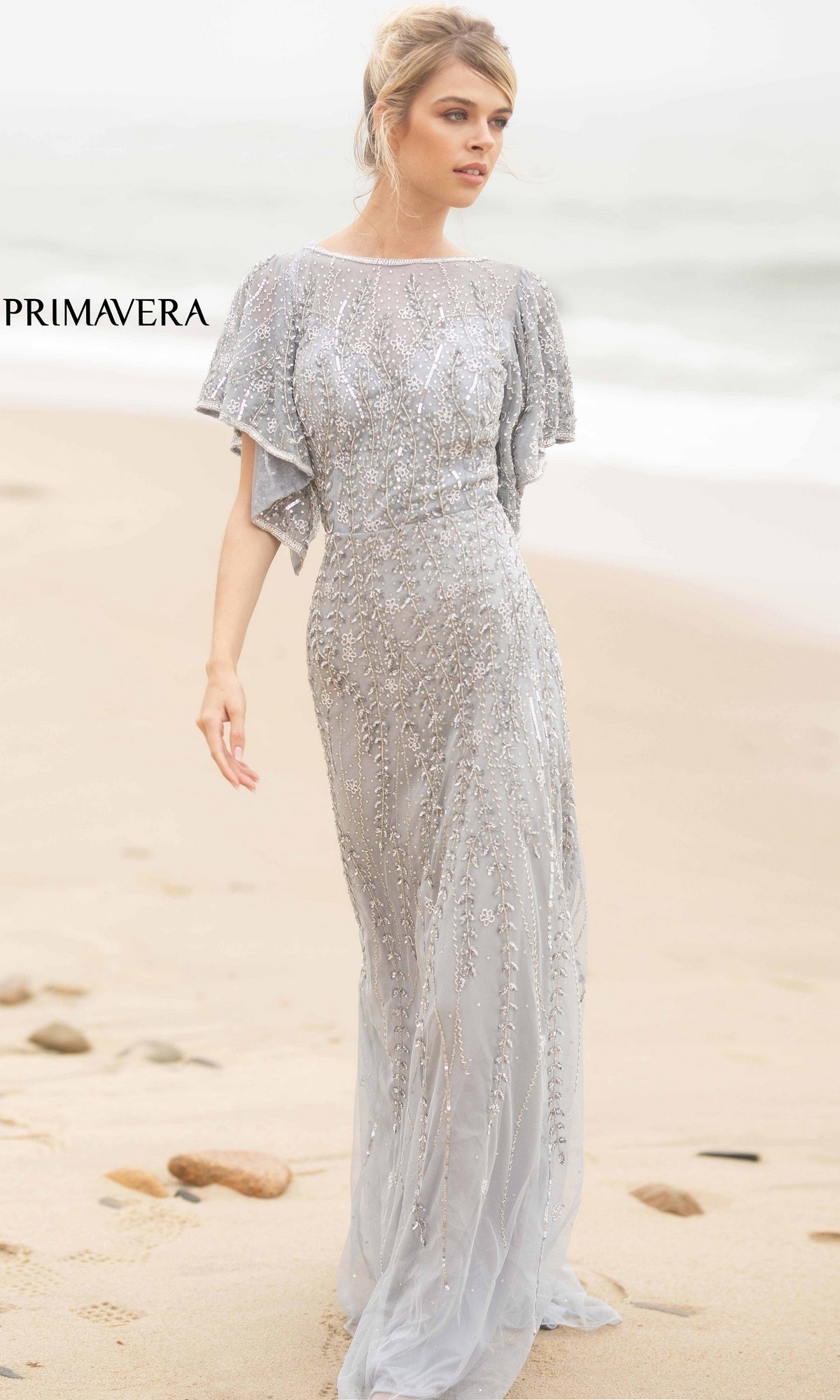A woman stands on a sandy beach with waves behind her, wearing the Short-Sleeve Beaded Long Prom Dress 3681—a silver, floor-length gown with flutter sleeves and intricate embellishments for an elegant evening look.